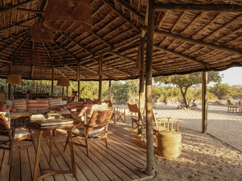 A luxurious open-air safari lodge lounge with a thatched roof and comfortable seating overlooking a sunny landscape.