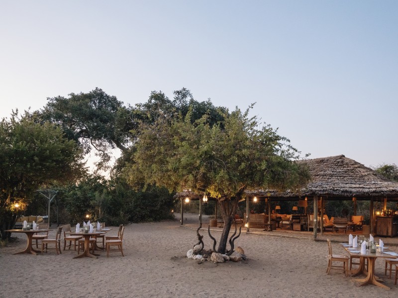 An elegant outdoor dining area at a safari lodge with thatched roofs and tables set on sand at twilight.