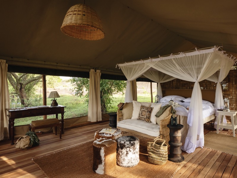 Interior of a luxury safari tent with a large canopy bed, mosquito netting, and wicker furniture overlooking a green savanna.