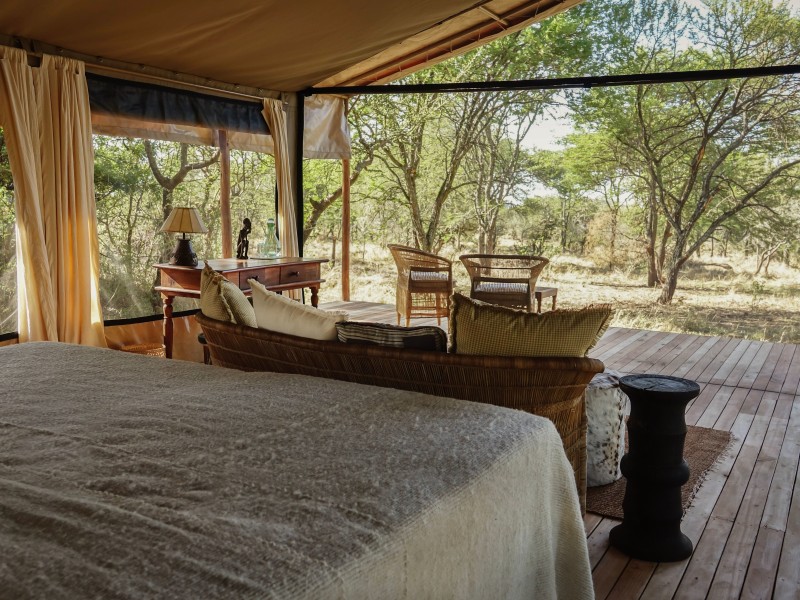 Inside a luxury safari tent with an open view of the African savanna, featuring a bed, a desk, and a lounge area leading to a wooden deck.