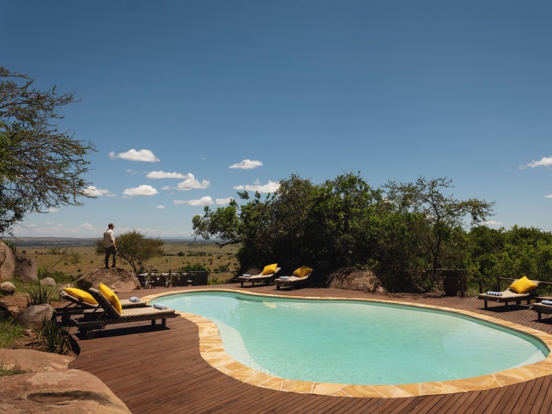 A kidney-shaped swimming pool on a dark wooden deck with yellow-cushioned lounge chairs, overlooking a vast savanna under a clear blue sky.