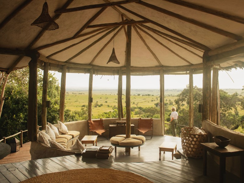 A high-end safari lodge interior featuring a thatched roof and comfortable seating, overlooking an expansive green savanna.