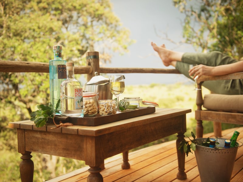 A variety of drinks and snacks on a wooden table on a deck overlooking a green landscape with a person relaxing.