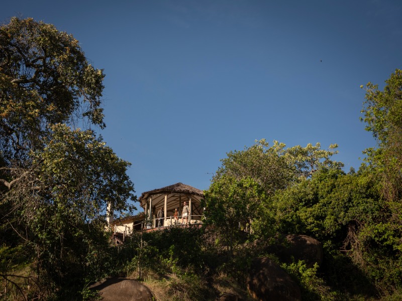 A rustic safari lodge with a thatched roof and balcony nestled among lush green trees and rocky terrain under a clear blue sky.