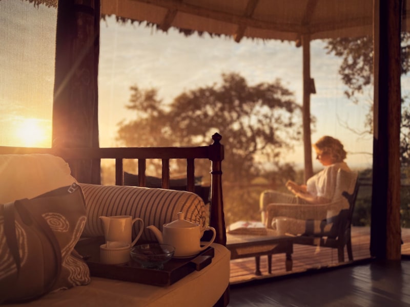 A tea set on a wooden tray in the foreground with a woman reading a book on a terrace at sunset.