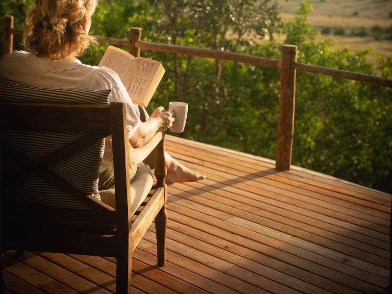 A woman sits on a wooden deck, reading a book and holding a mug while overlooking a vast landscape at sunset.