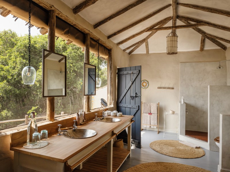 Rustic safari lodge bathroom featuring a double wooden vanity with metal sinks and large windows overlooking lush trees.