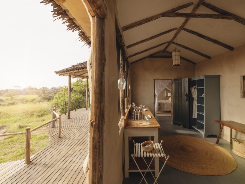 Interior of a luxury safari lodge with earth-colored walls and a wooden deck overlooking a golden savannah landscape.