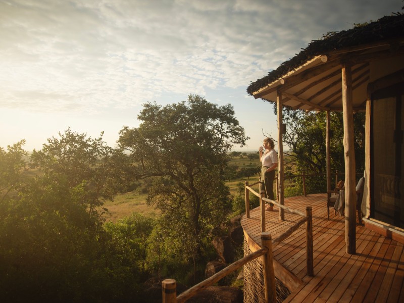 A woman stands on a rustic wooden deck of a safari lodge at sunset, overlooking a vast African landscape.