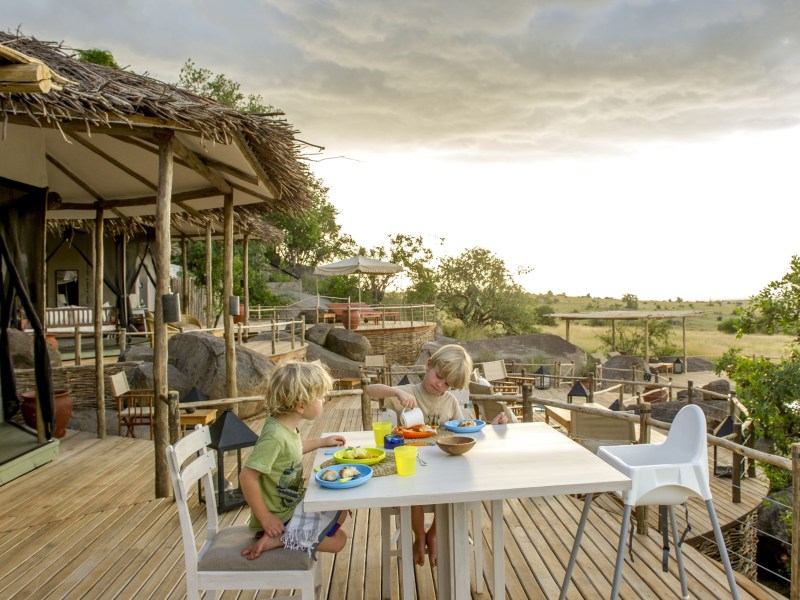 Two young boys enjoying a meal at an outdoor table on a wooden deck overlooking a vast savanna at a safari camp.