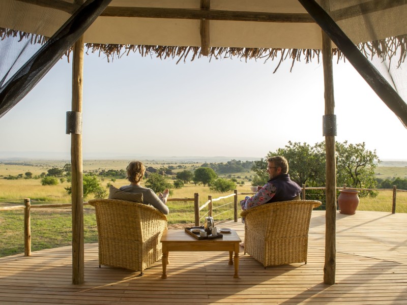 Two people sit in wicker chairs on a wooden deck, looking out over a vast savanna landscape under a soft morning sun.