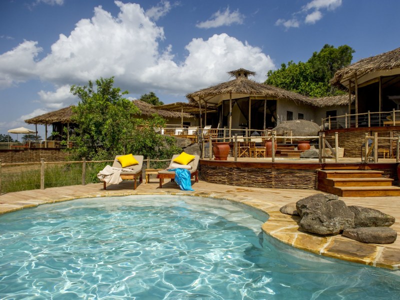 A luxury safari lodge featuring a crystal clear swimming pool and thatched-roof buildings under a blue sky.