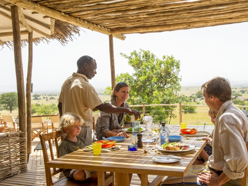 A family enjoying an outdoor lunch at a safari lodge while a waiter serves wine, overlooking a vast savanna landscape.