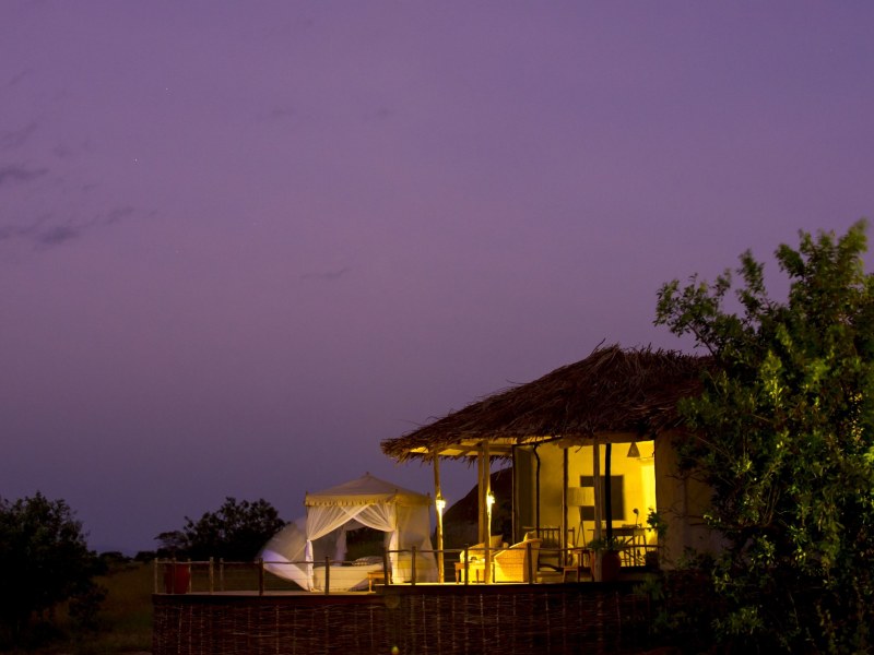 A luxury safari lodge with a thatched roof and an outdoor canopied bed glowing warmly against a purple twilight sky.
