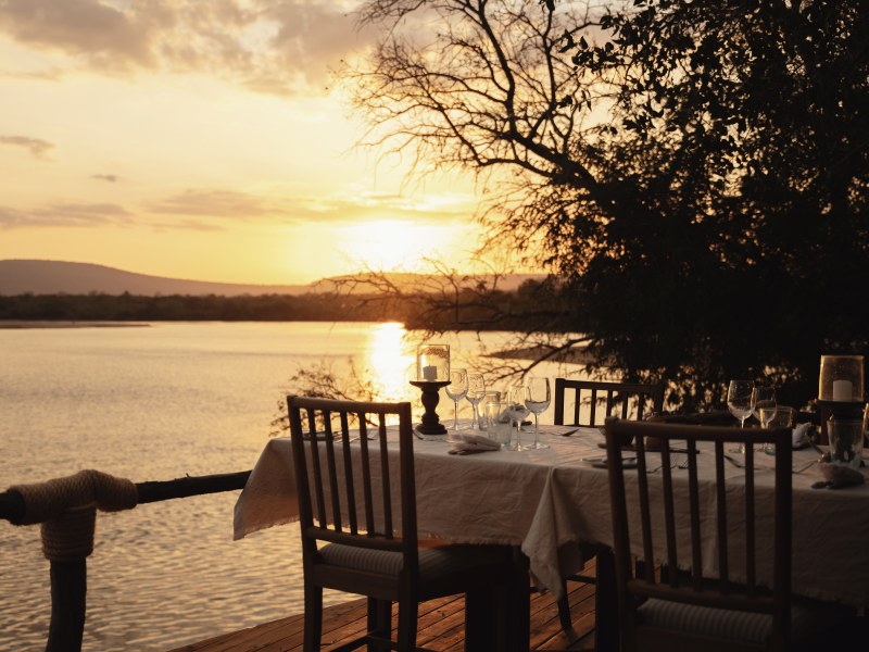 A formal dining table set on a wooden deck overlooking a river during a golden sunset.