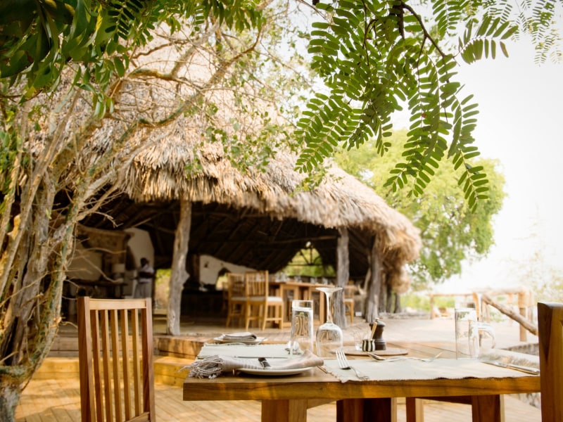 A set dining table at an outdoor restaurant featuring wooden furniture and a large thatched roof structure in the background.