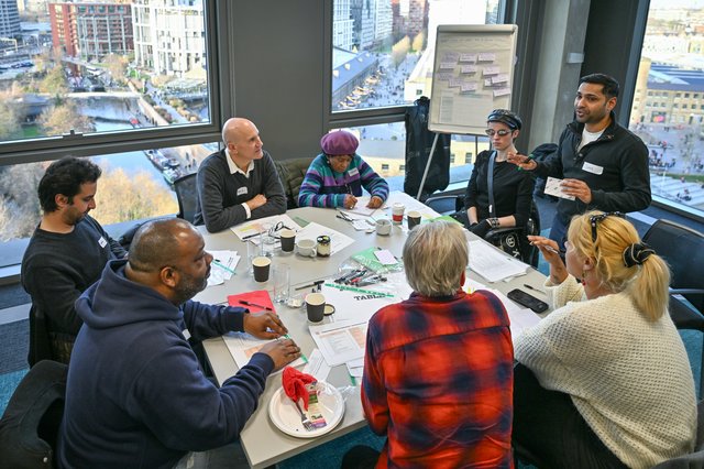 Seven panel members sat round a table. There is a facilitator stood speaking. A view of Camden out the windows behind and a flip chart board behind the table with post its on.