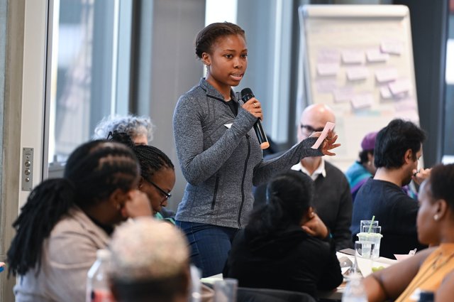 A panel member is stood in a grey jacket with a microphone speaking to a room of other panel members. There is a blurry flip chart with post its in the background.