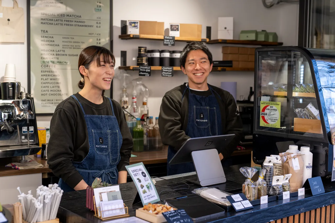 Two team members smile behind the serving desk next to snacks and cakes for sale.