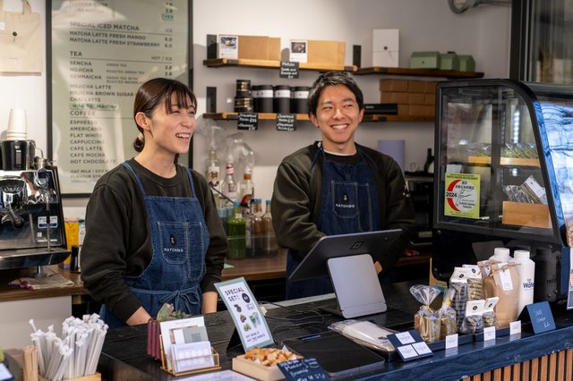Two Matchado team members smiling behind the serving desk at their shop with merchandise behind them and snacks on show to the right of them.