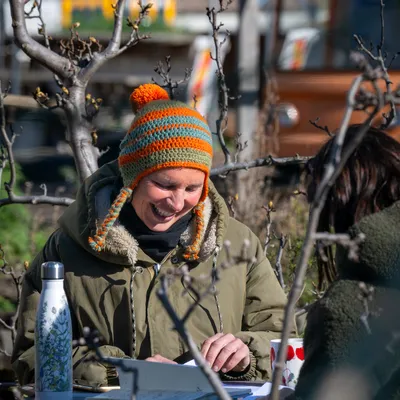 A smiling team member story garden sits behind a table in between trees with a bright orange and green hat on.