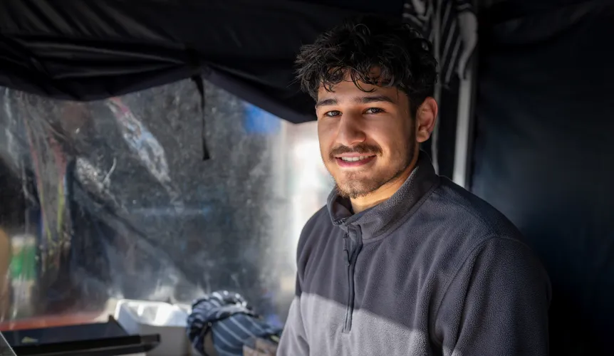 Market stall team member stands smiling in a fleece stands in front of a grill and towels inside a dark coloured marquee.