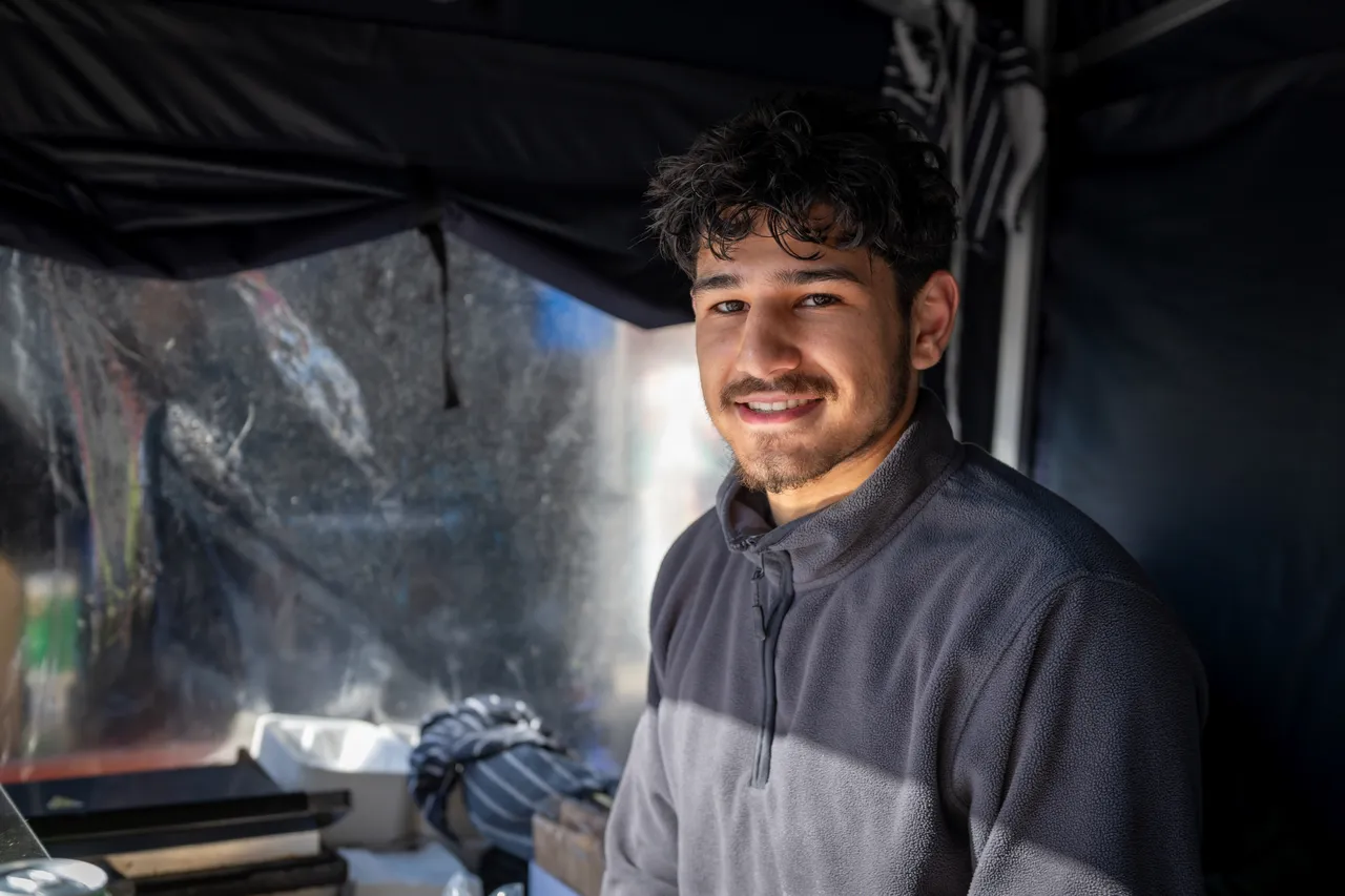 Market stall team member stands smiling in a fleece stands in front of a grill and towels inside a dark coloured marquee.