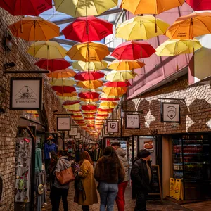 Red, orange, yellow and pink umbrellas in Camden Market above the shop fronts and signs in the Camden market passageway. People mingle in front of the shops.