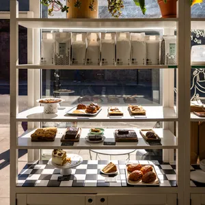 A white bakery display cupboard stands in front of a window with cakes and pastries on it. There is a road with blue sky above it outside the window.