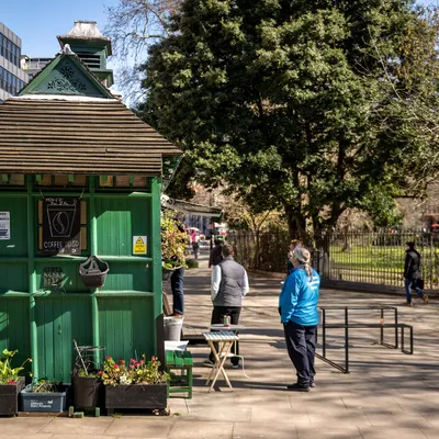 Green, wooden, small coffee shop next to a park with people stood around.