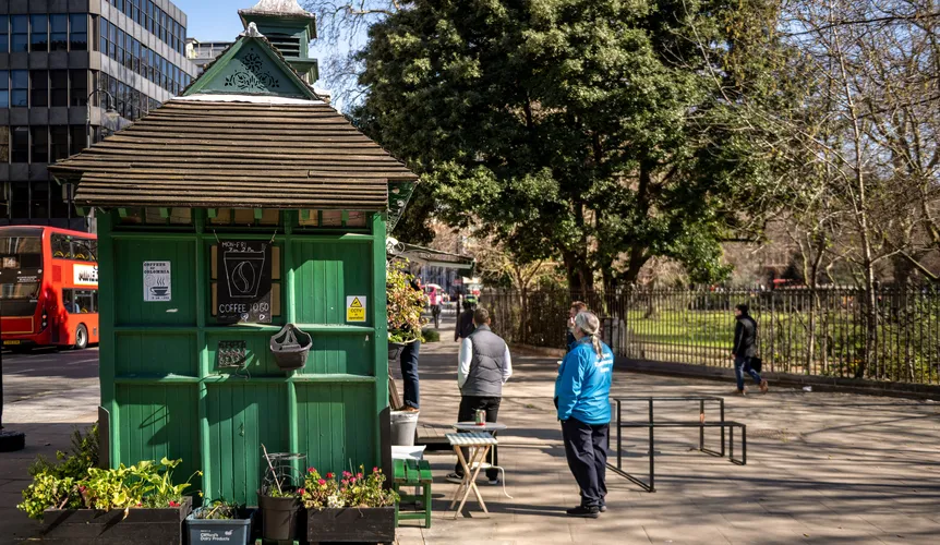 Green, wooden, small coffee shop next to a park with people stood around.