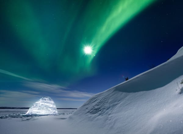 Small glowing igloo at base of snow drift under vivid green aurora with moon