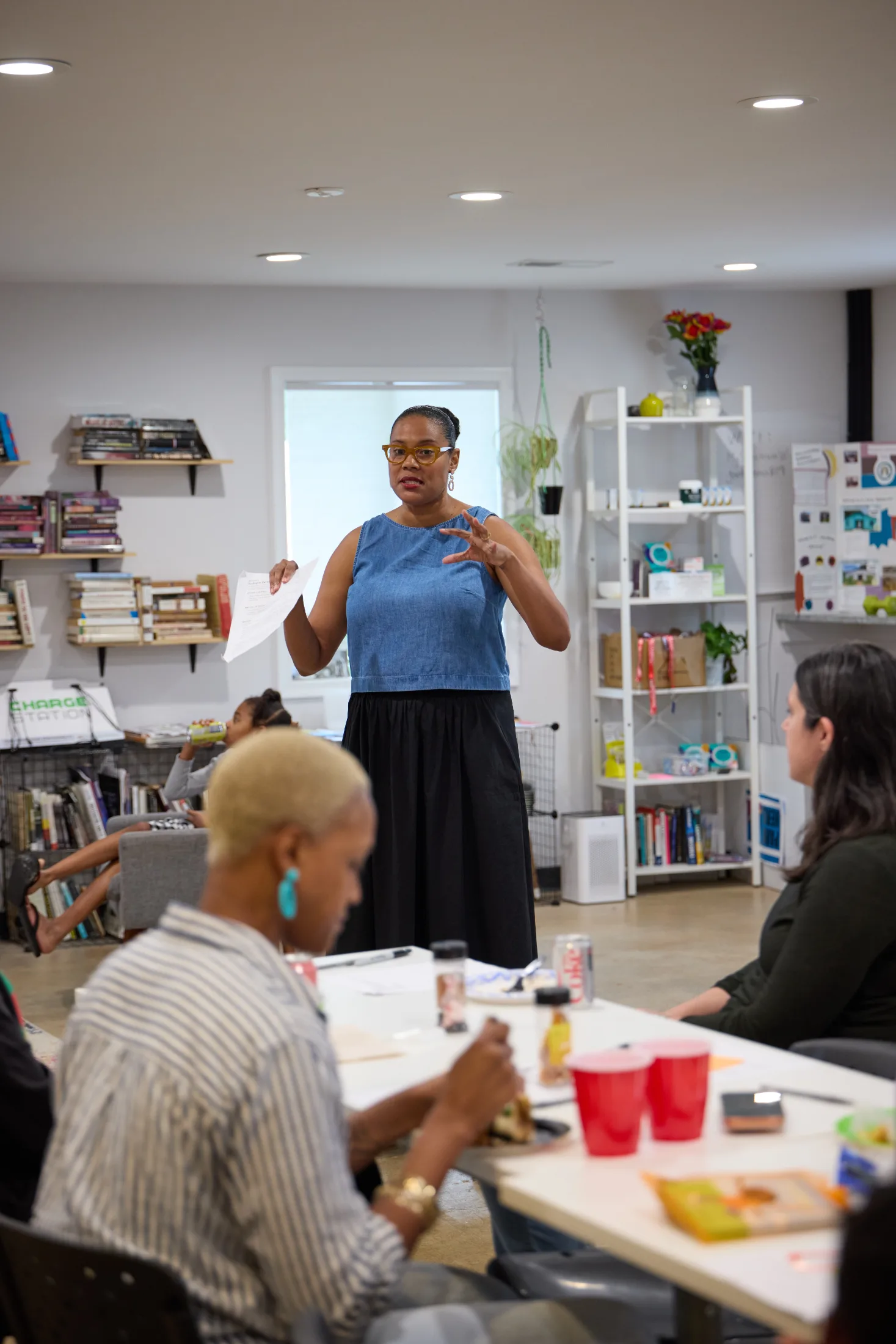 Tiffany, founder of Bright Black, stands holding a sheet of paper and gesturing as she speaks to participants seated around a table. She wears glasses, a blue sleeveless top, and a black skirt. Bookshelves, plants, and product displays fill the community space behind her.