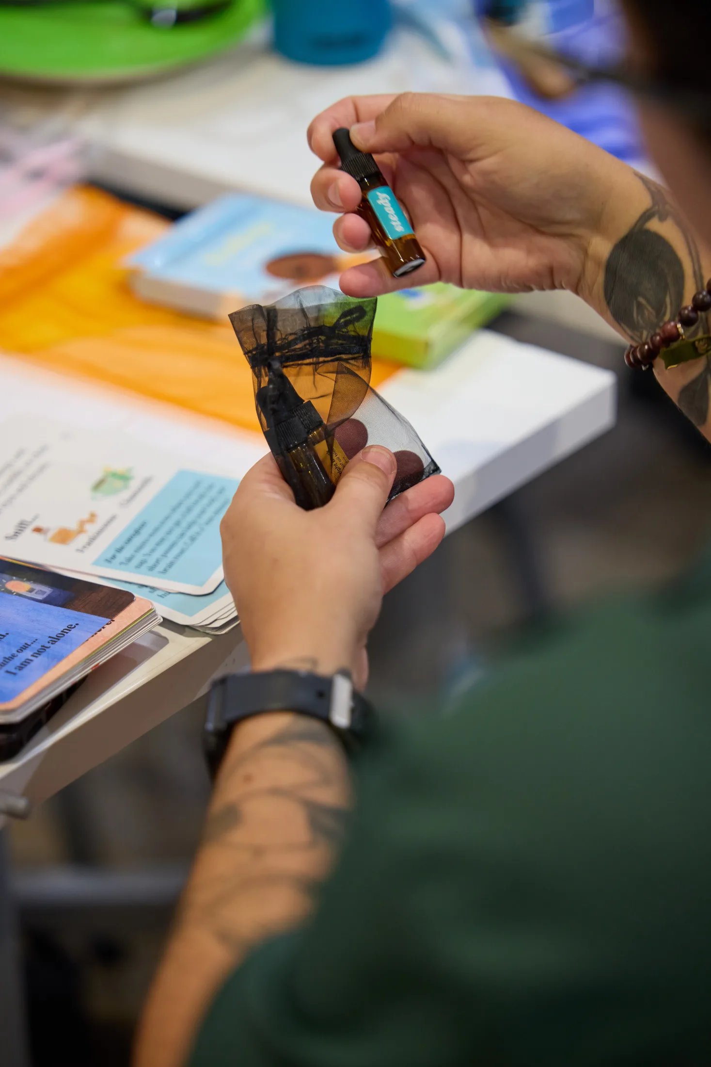 Close-up of a workshop participant holding a small amber bottle of scented oil labeled “Happy” in one hand and a black mesh pouch containing additional bottles in the other. Colorful activity cards and workshop materials are spread across the table.