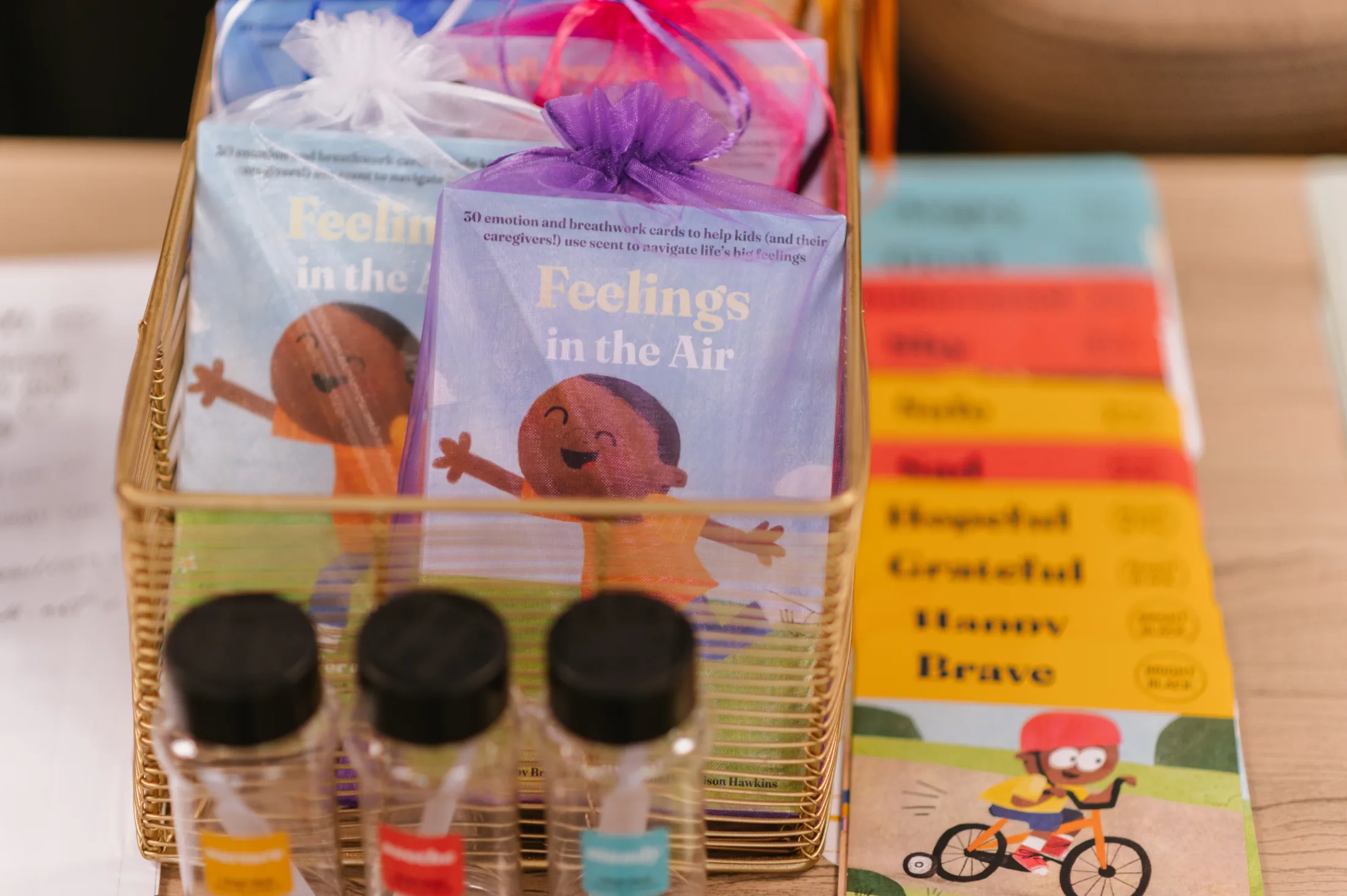 Colorful Feelings in the Air activity card sets featuring illustrated children are displayed in a gold wire basket alongside small scent bottles. The kits include emotion and breathwork cards designed to help children and caregivers navigate big feelings.