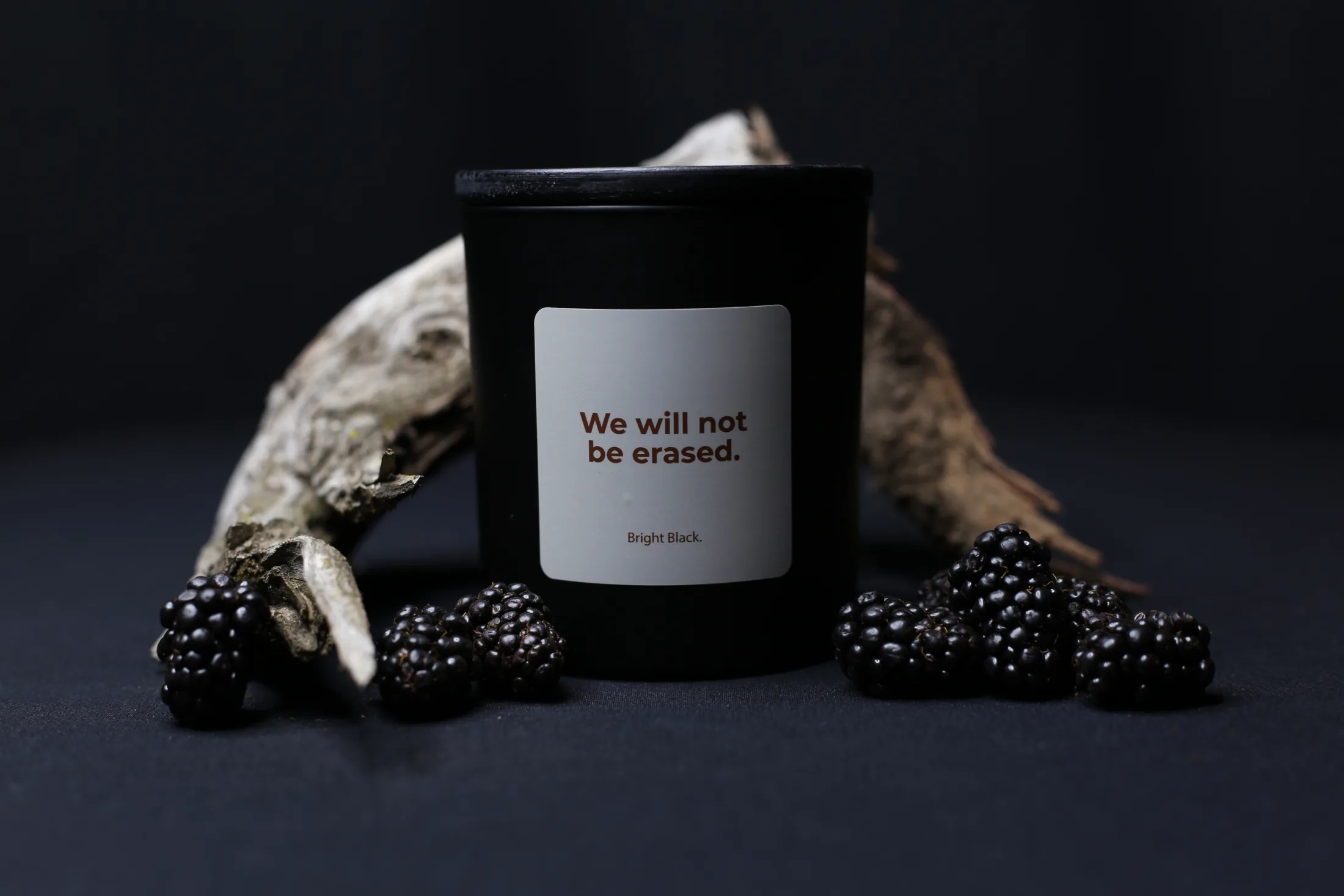 A black Bright Black candle with a white label reading “We will not be erased” sits against a dark background, surrounded by fresh blackberries and pieces of weathered wood.