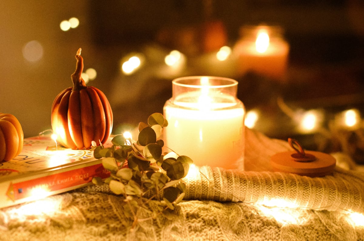 A lit candle, small pumpkins and eucalyptus on a knit blanket with fairy lights