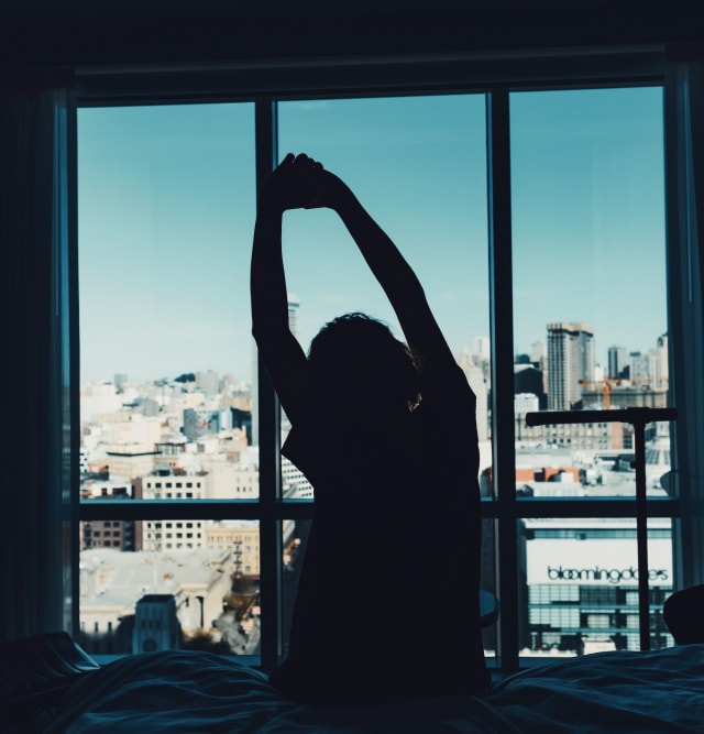 A person sitting in bed stretching both arms overhead in front of a large window with a city view
