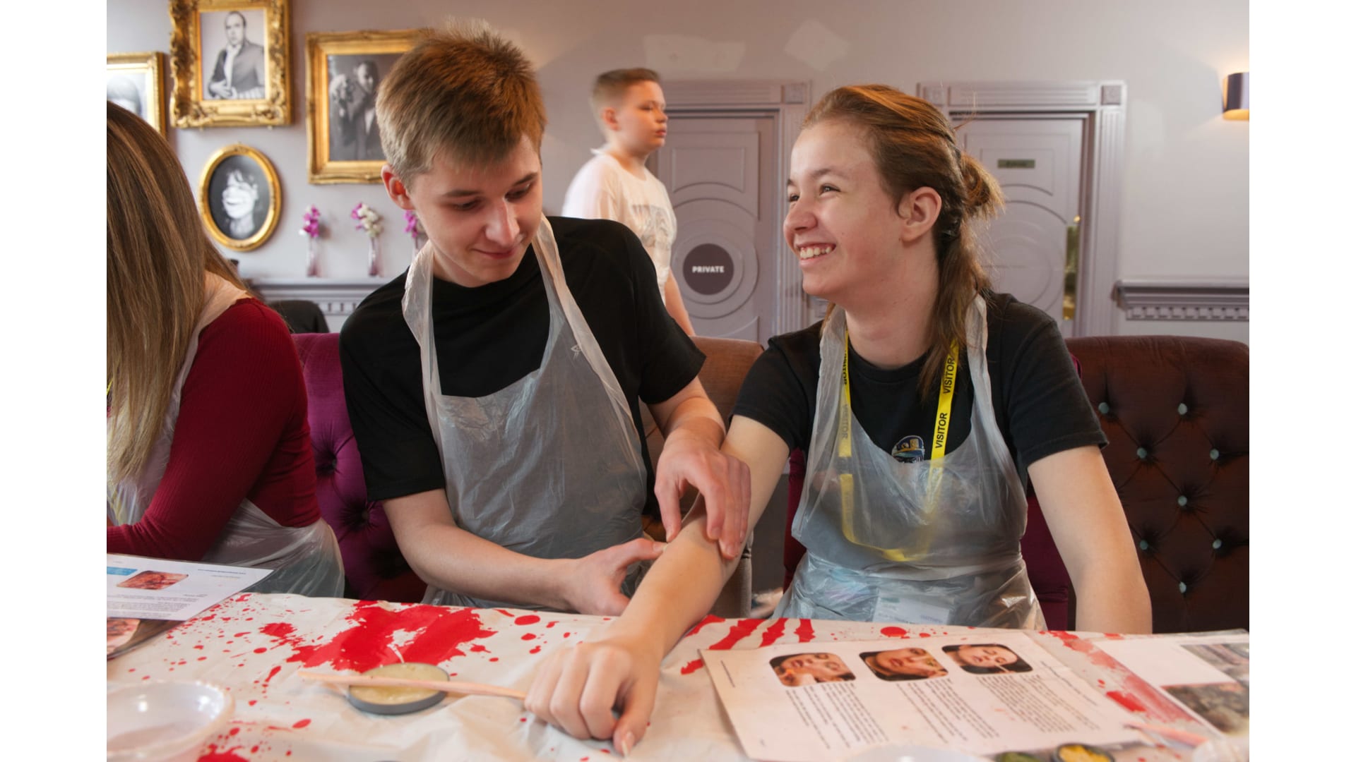 Two young people wearing clear plastic aprons sit at a table splattered with red stage-blood paint; one applies make-up to the other's forearm using a small brush, with reference sheets visible on the table