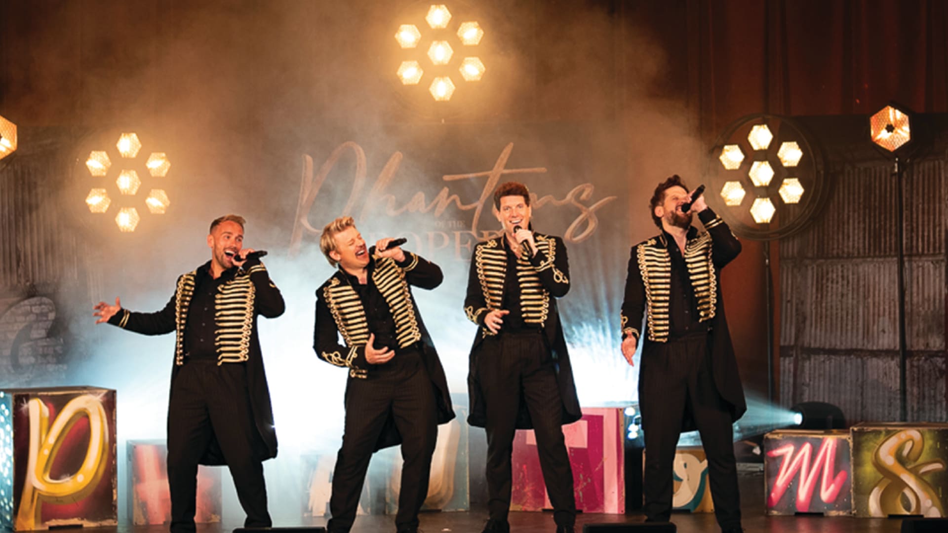Four male performers on a fog-filled stage in matching black and gold military-style jackets holding microphones, with the show title Phantoms of the Popera on the backdrop.