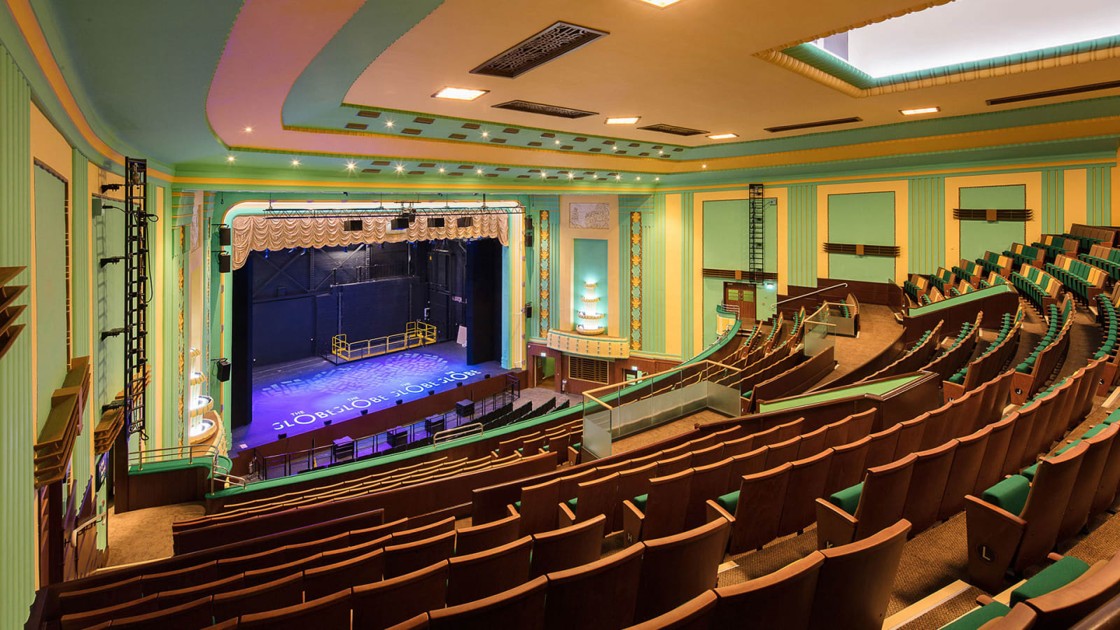 Interior of Stockton Globe's auditorium showing curved rows of brown and green seats facing a proscenium stage lit in violet and blue, with art deco gold and green wall detailing.