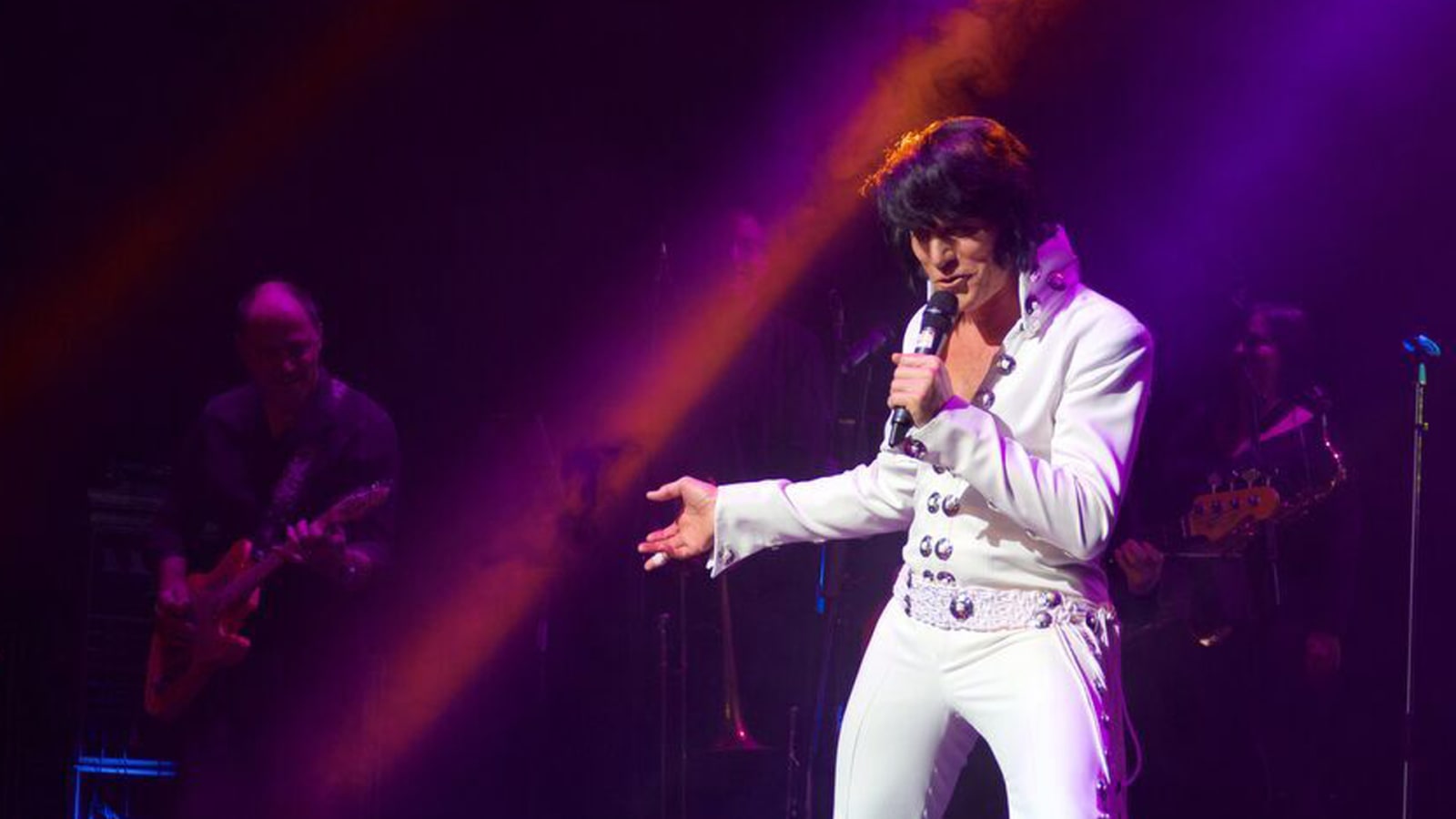 A performer in a white jumpsuit sings into a microphone on a darkened stage with purple and orange lighting.