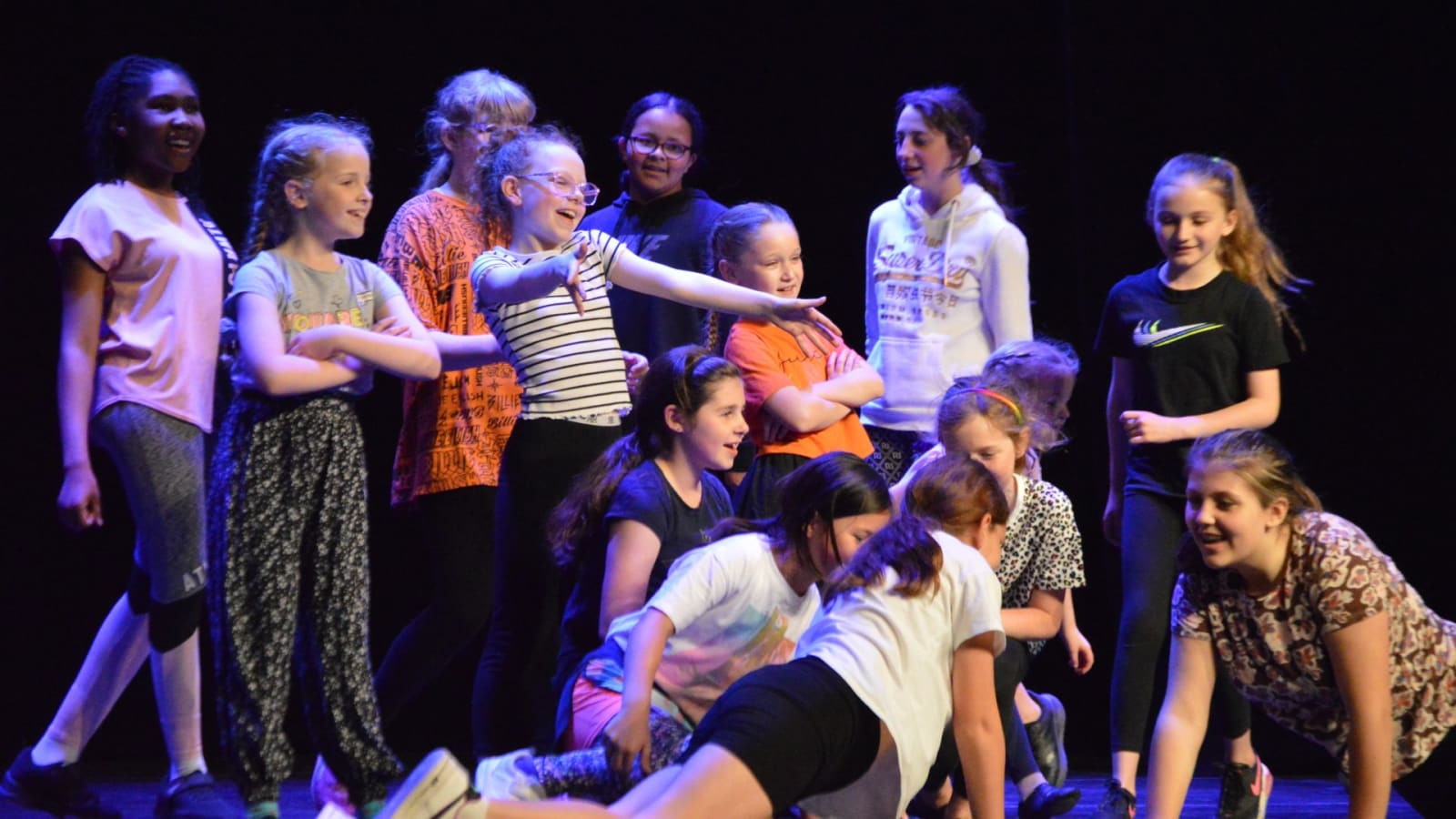 A group of children on a darkened stage lit with purple and warm light, one child standing centre with an arm outstretched while others are arranged around her in various poses.