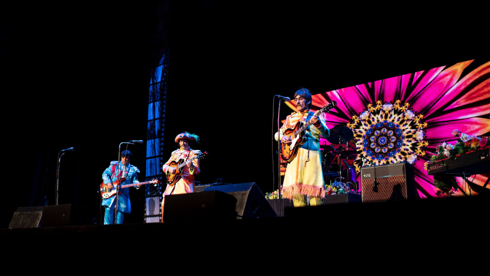 Three guitarists in colourful Sgt Pepper-style costumes perform on a darkened stage beside a large LED screen displaying a vivid pink and yellow mandala pattern, with a Vox amplifier visible to the right.