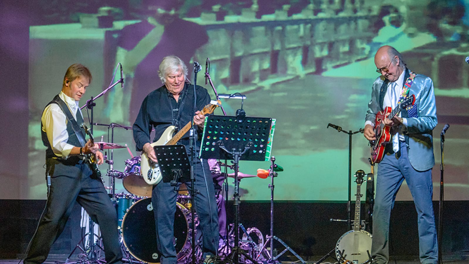 Three guitarists performing on a lit stage, a drum kit behind them and a projected black-and-white street scene on the backdrop; one wears a light blue jacket, another all black.