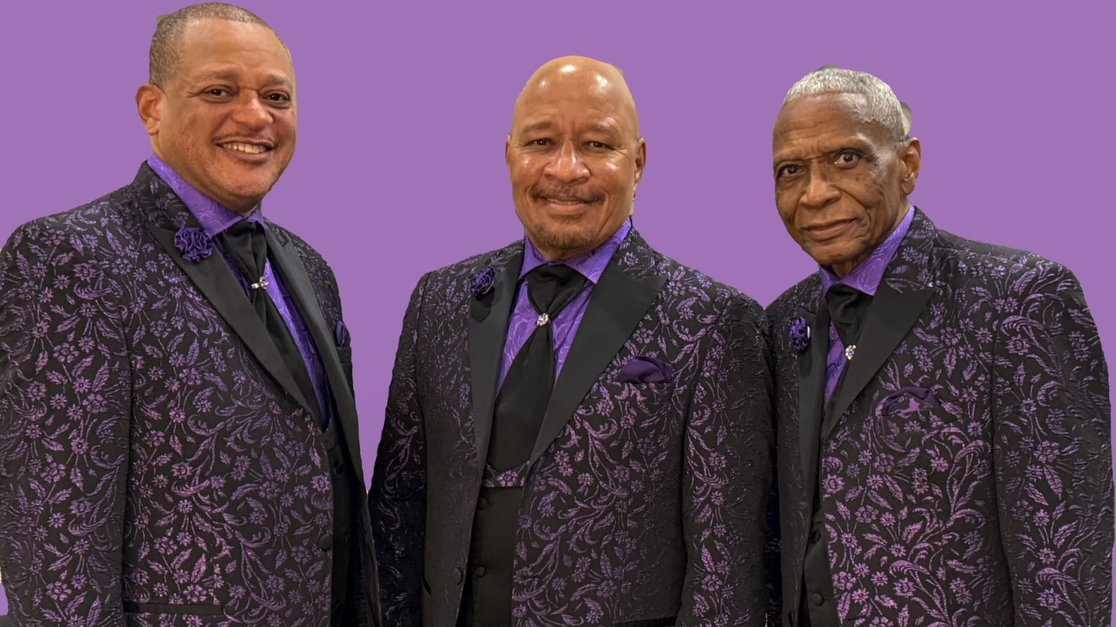 Three men in matching black and purple floral jackets with purple shirts and black ties, posed against a solid lavender background