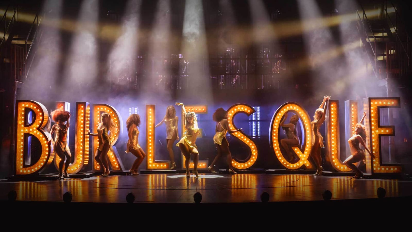 Performers in gold and amber costumes pose and dance in front of large illuminated marquee letters spelling BURLESQUE on a stage lit with white spot beams and purple haze.