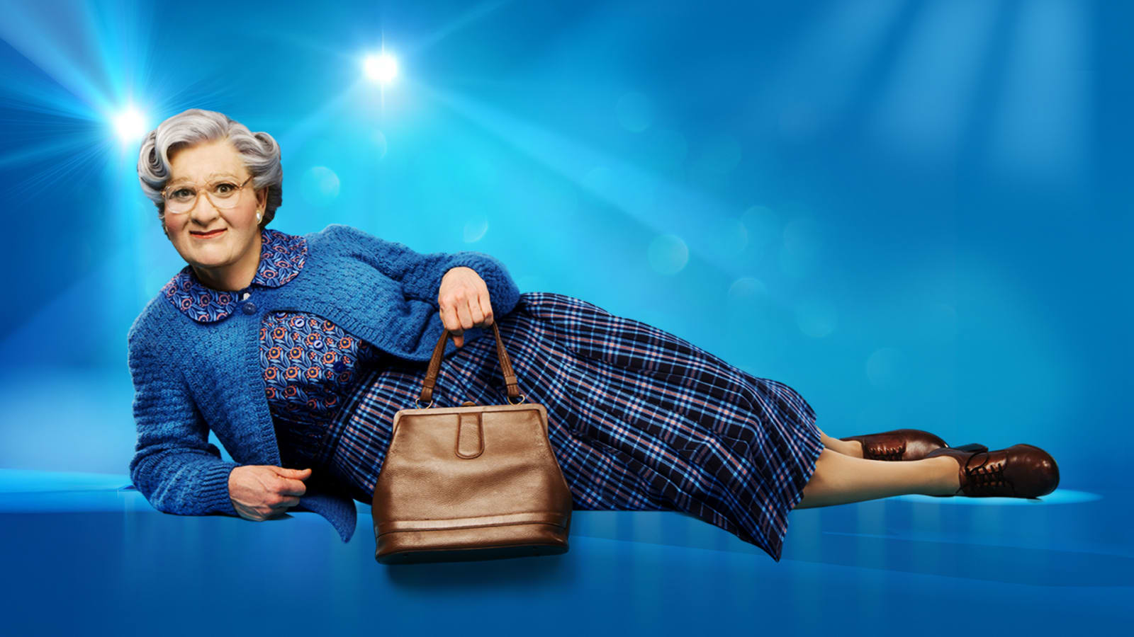 A performer in Mrs. Doubtfire costume with grey curled wig, round glasses, blue cardigan and tartan skirt reclines holding a tan handbag against a bright blue background.
