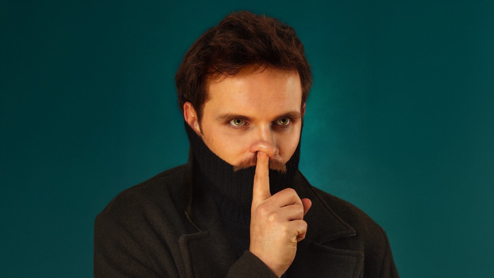 A man in a black coat and roll-neck raises one finger to his lips against a teal background.