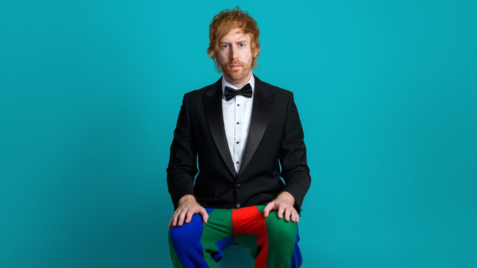 A person with short auburn hair and a beard sits wearing a black tuxedo jacket with bow tie and multicoloured patchwork trousers in blue, green and red, against a teal background.